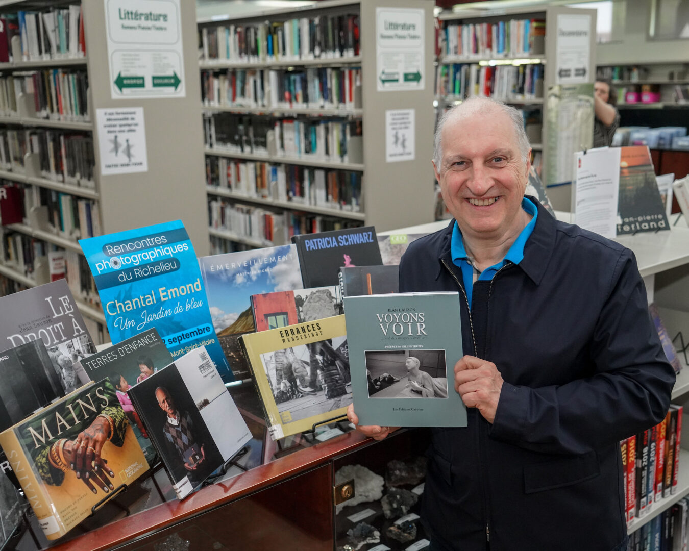 Voyons voir est le 26e livre publié par les Éditions Cayenne de Mont-Saint-Hilaire, auquel a notamment participé son éditeur Robert Hébert, qu’on reconnaît à la bibliothèque Armand-Cardinal devant quelques-uns des autres livres qu’il a édités. Photo François Larivière | L’Œil Régional ©