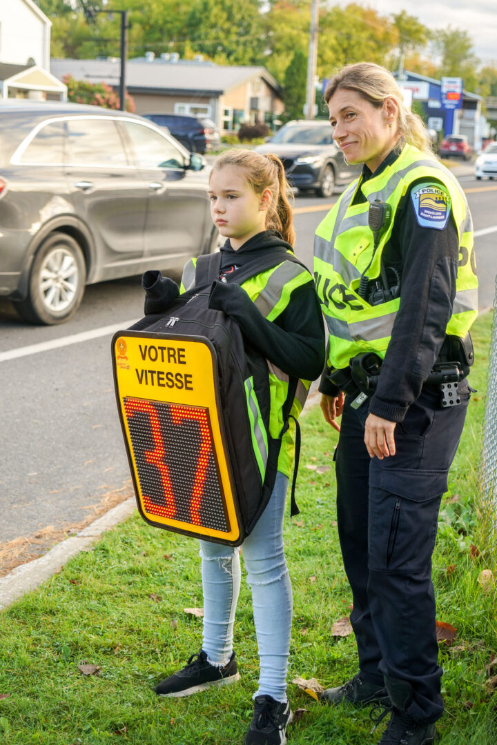 Anaïs Larouche porte un sac à dos muni d'un afficheur de vitesse pour sensibiliser les automobilistes. Elle est accompagnée de la policière Sonia Villiard.
Photo François Larivière | L’Œil Régional ©
